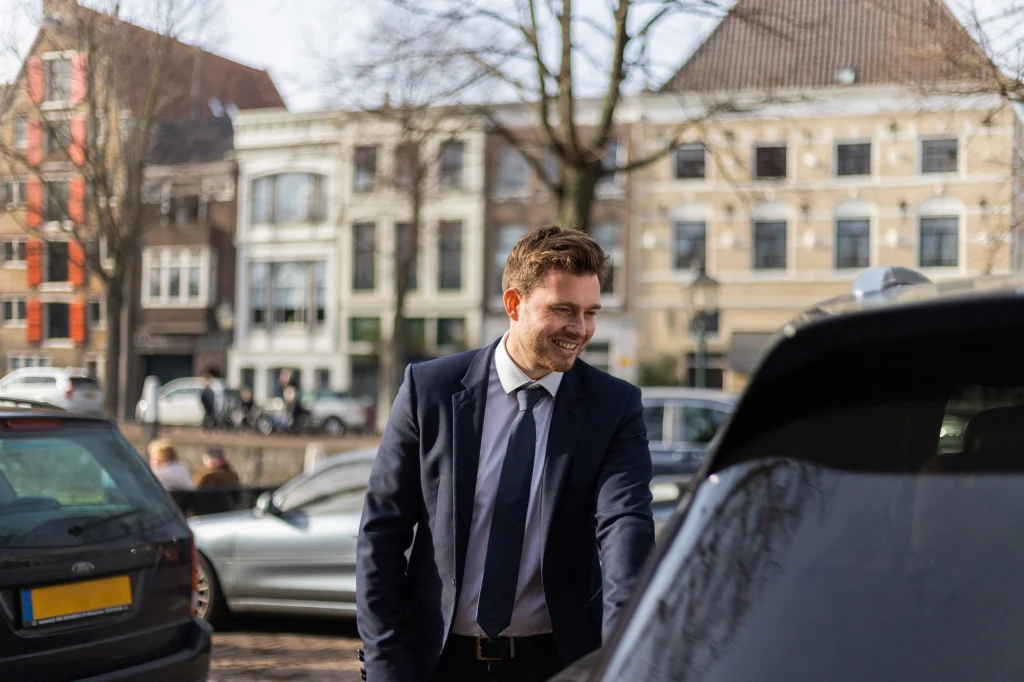 Glimlachende chauffeur van Taxi Stam in pak naast een auto in de stad
