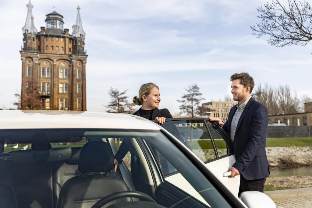 Chauffeur en passagier bij een witte auto met de watertoren op de achtergrond