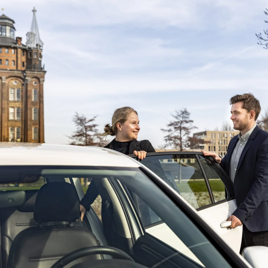 Chauffeur en passagier bij een witte auto met de watertoren op de achtergrond